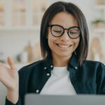 Smiling woman in glasses waving hand hello, answers video call, chatting online on laptop at home